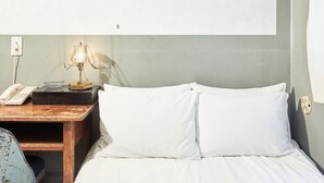 Uma cama bem arrumada com lençóis brancos, uma mesa de cabeceira com abajur, telefone e um pequeno objeto decorativo.