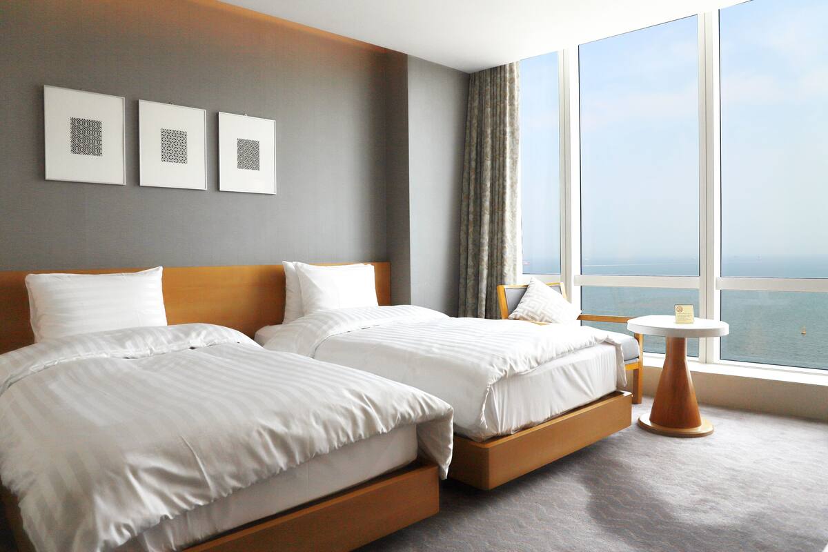 A hotel room with two beds, a small table, and a large window with a view of the sea.