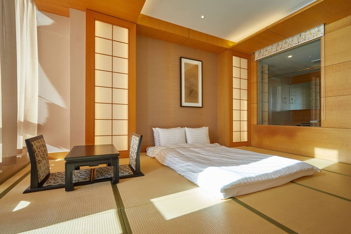 A traditional Japanese-style bedroom with tatami flooring, a bed, a small table with a chair, and a sliding glass door leading to a bathroom.