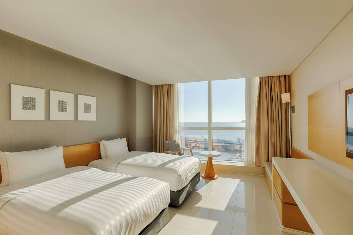 A hotel room with two beds, a large window with a city view, and a small table with a lamp.