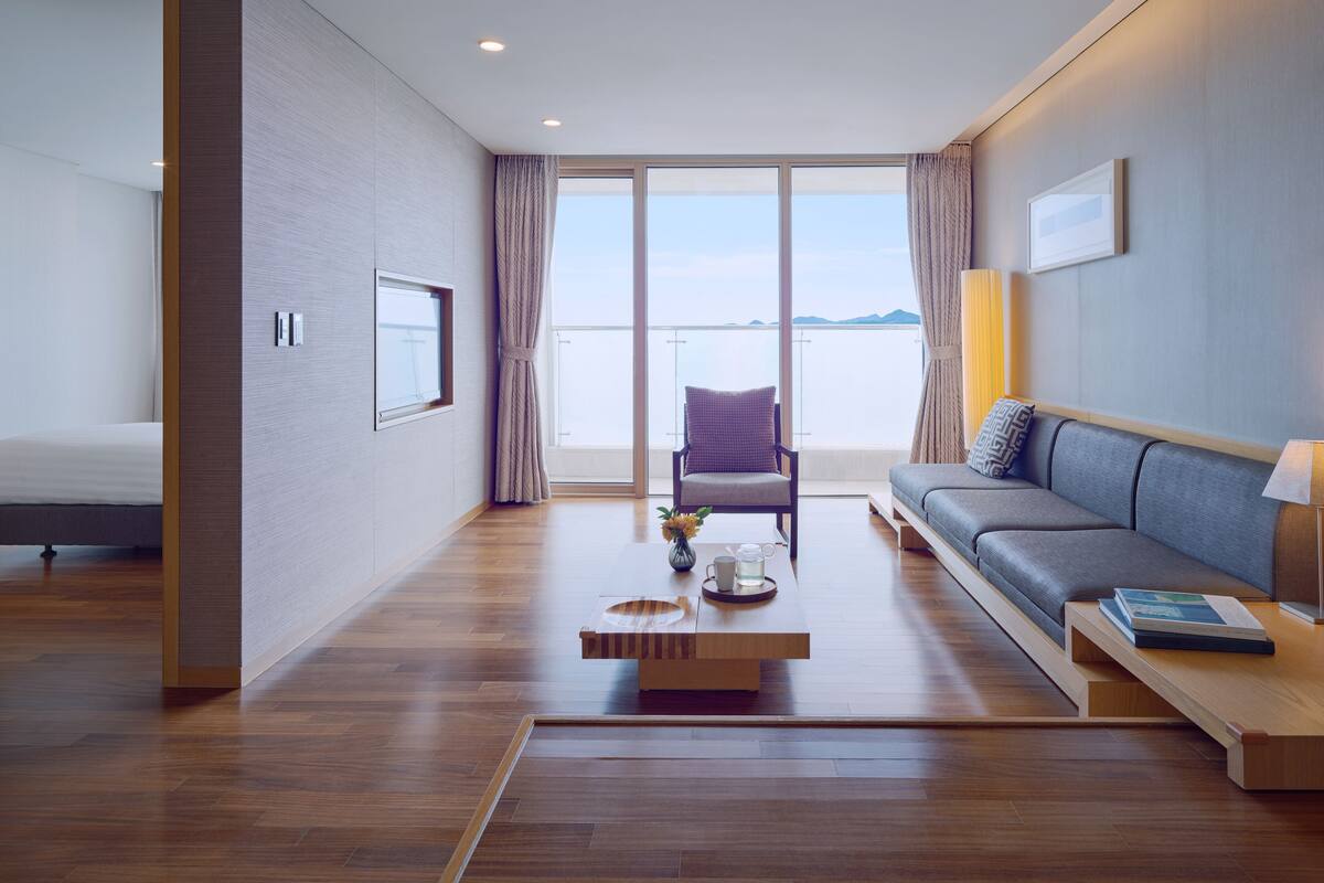 A modern hotel room with a large window, a sofa, a chair, and a wooden coffee table.