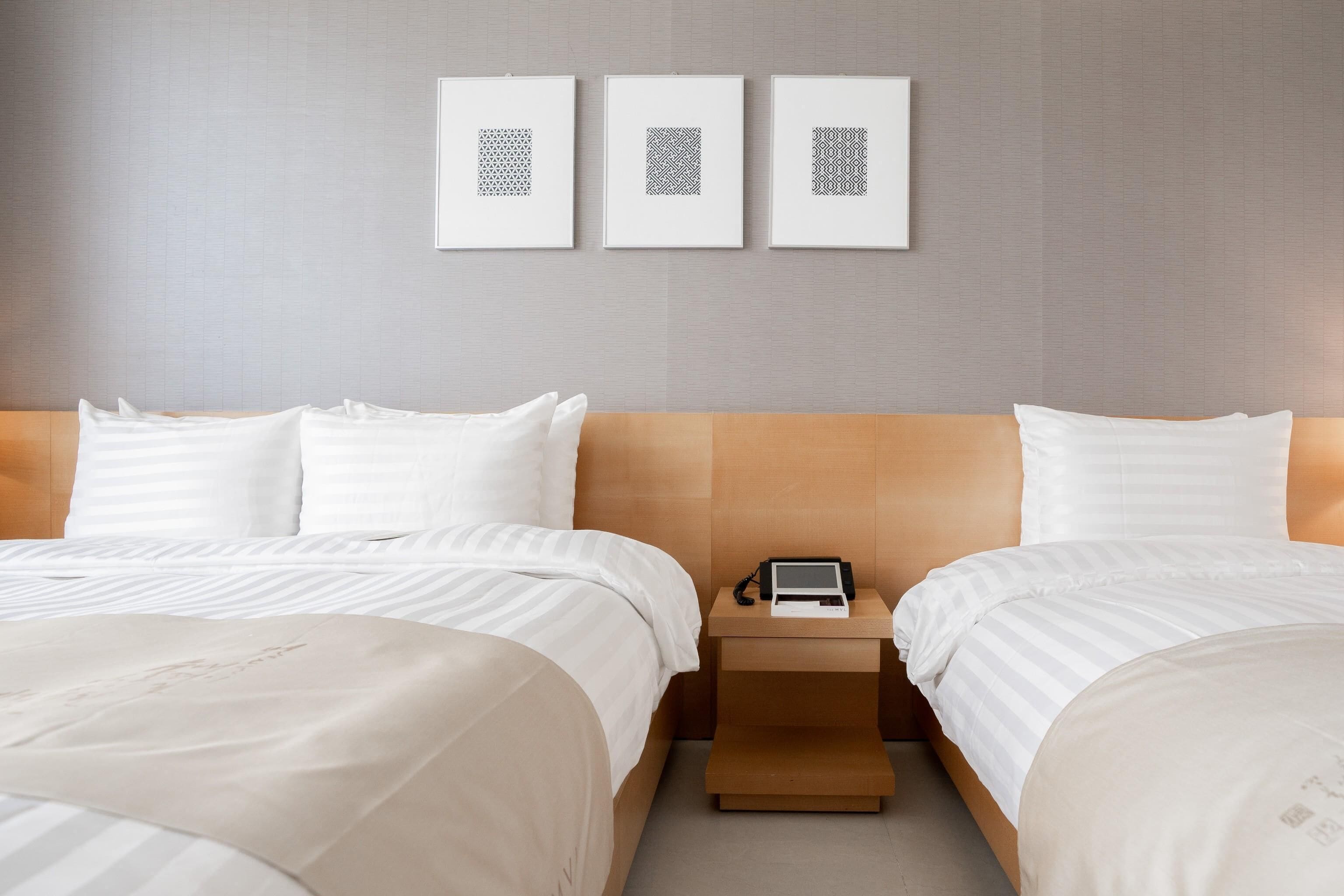 A hotel room with two beds, a nightstand with a phone and a tablet, and three framed wall art pieces.