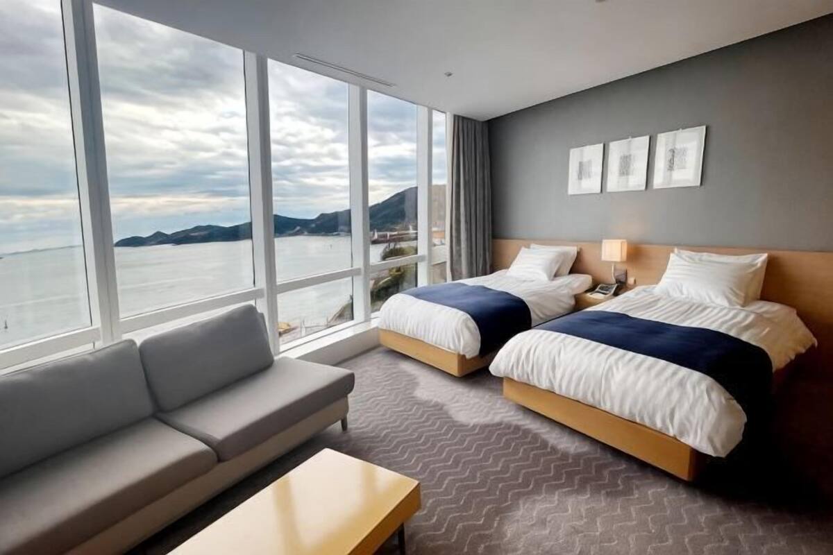 A hotel room with two beds, a sofa, a small table, and large windows with a view of the sea and mountains.