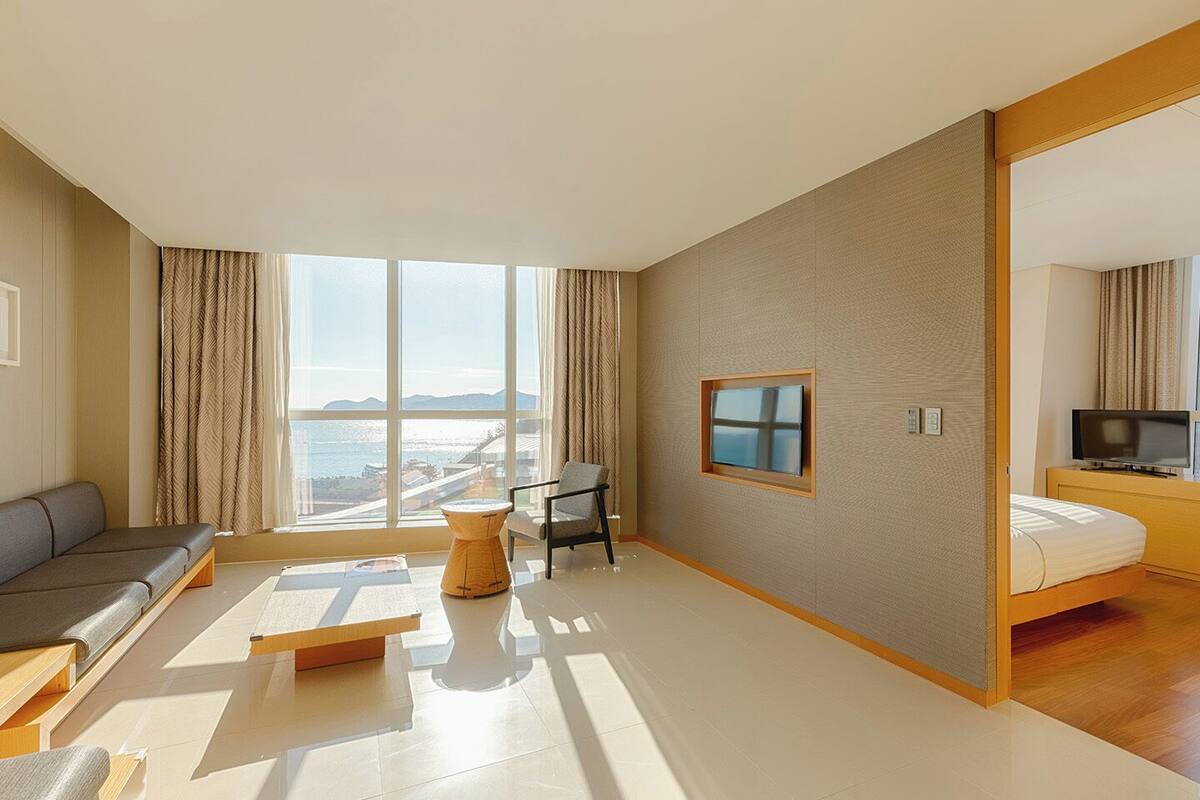 A modern hotel room with a large window, a sofa, a small table, a TV, and a bed.
