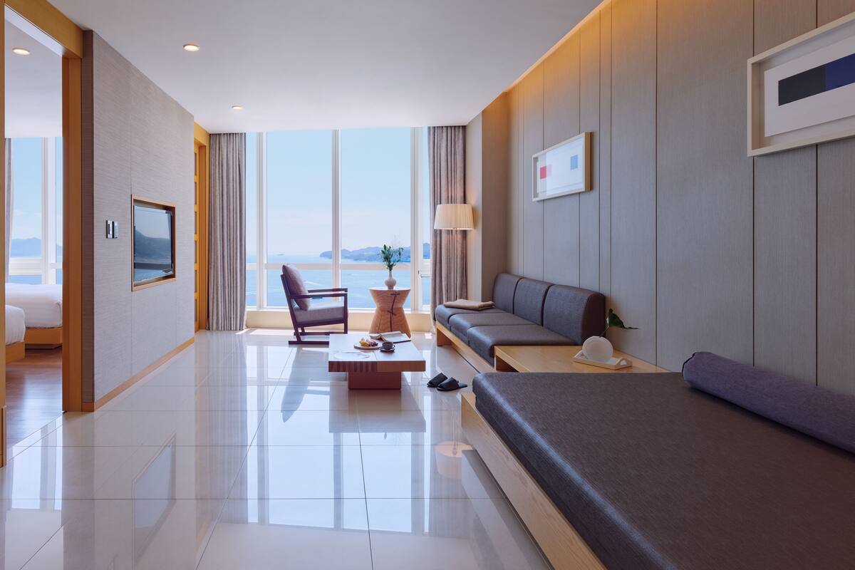 A modern hotel room with a large window, a sofa, a chair, a small table, and a bed.