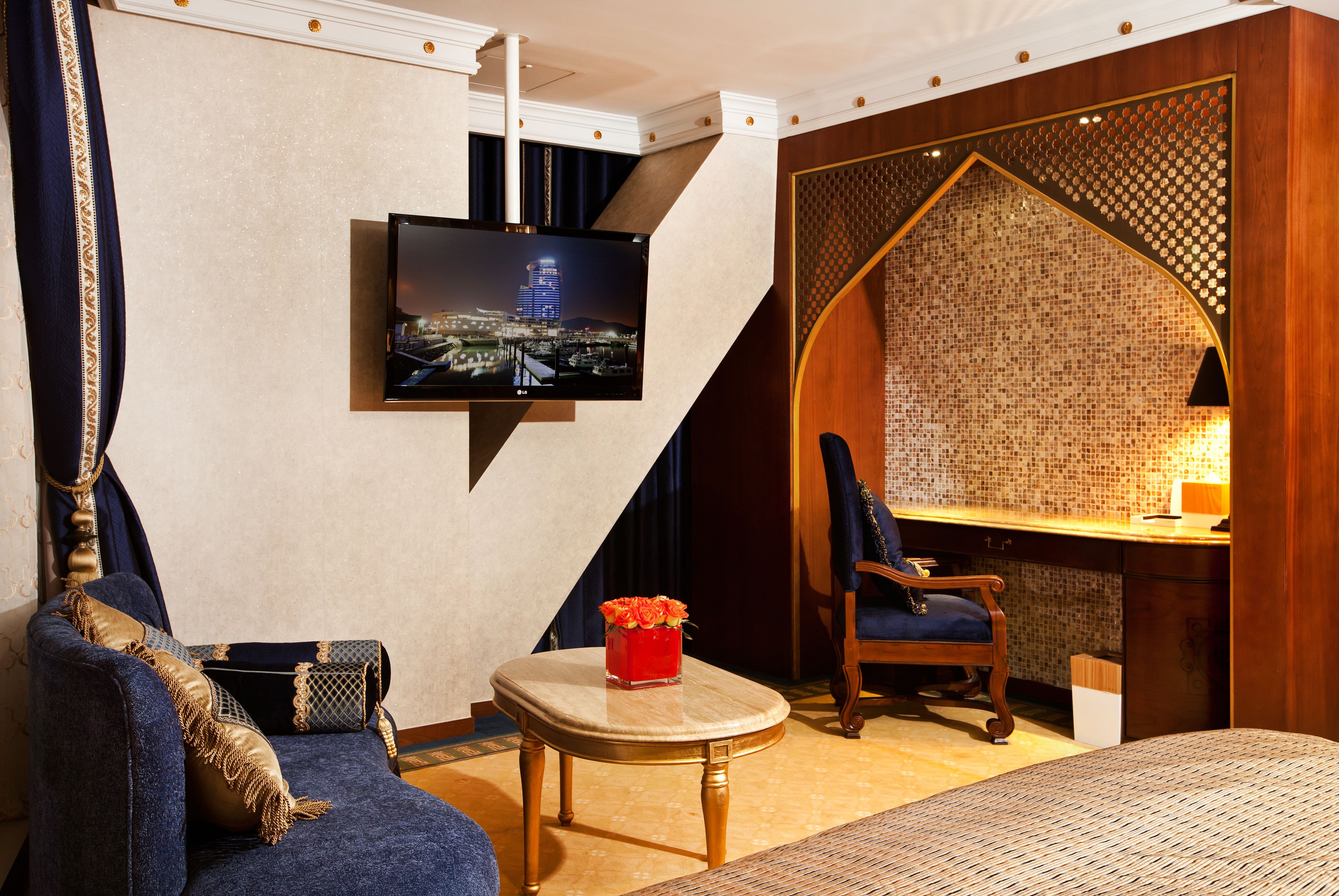 A hotel room with a large bed, a sofa, a small table with a vase of flowers, a desk with a lamp, and a television mounted on the wall.