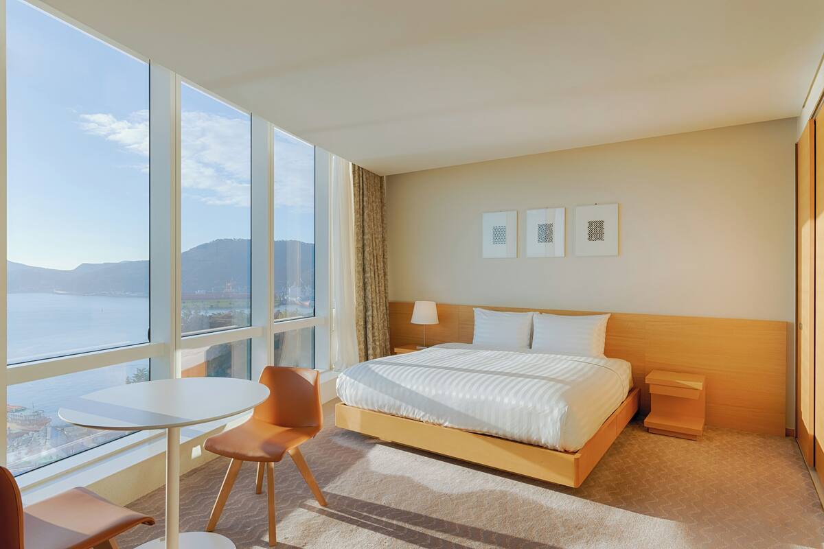 A hotel room with a large bed, a small table, a chair, and a view of the sea.