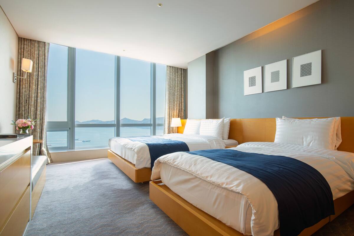 A hotel room with two beds, large windows, a view of the sea, and three framed pictures on the wall.