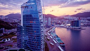 A modern skyscraper with a unique angular design, surrounded by a waterfront area with boats and docks. - Sono Calm Yeosu (Yeosu)