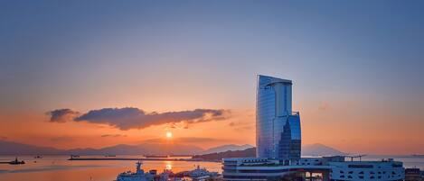 Un edificio moderno con un diseño arquitectónico único, ubicado cerca de una masa de agua con un atardecer de fondo.