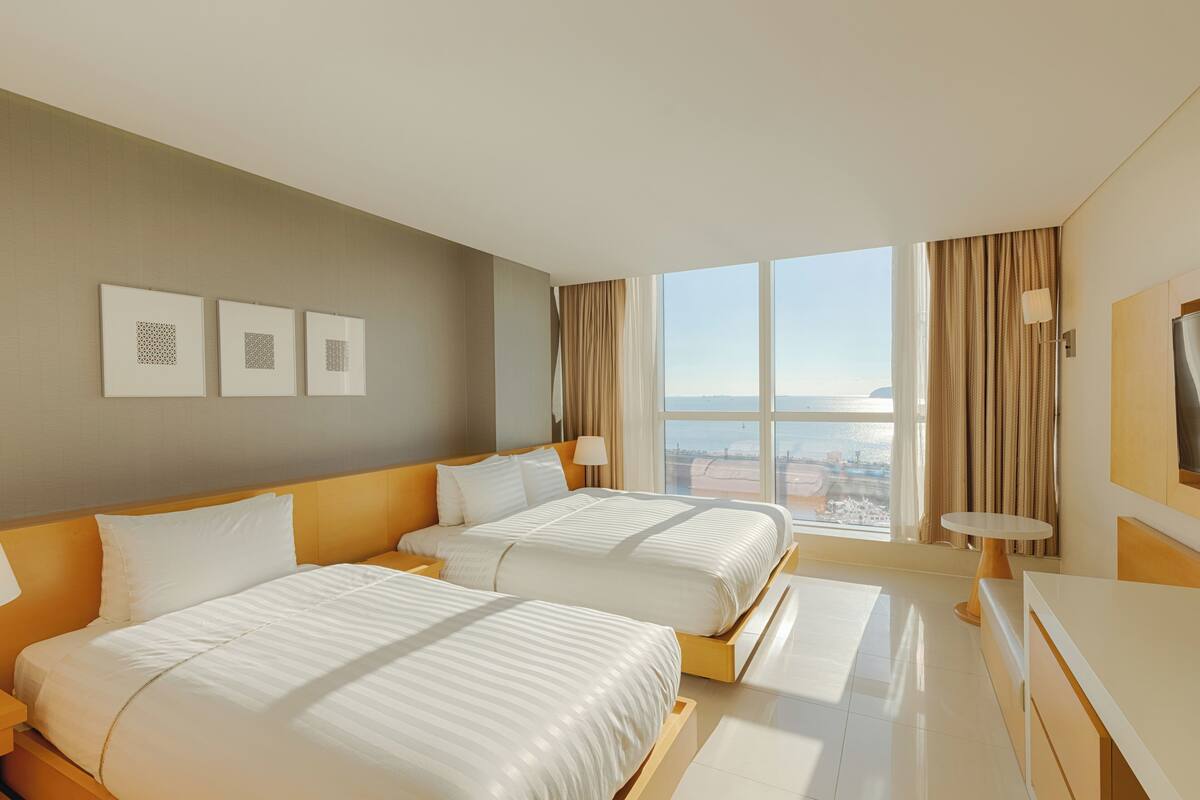 A hotel room with two beds, a large window with a city view, a small round table, and a flat-screen TV.