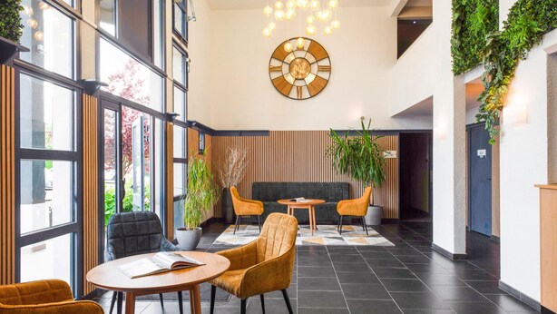 A modern lobby with large windows, a round wooden clock, and a seating area with orange chairs and a black sofa. - Kyriad Dieppe - Saint Aubin Sur Scie (Saint-Aubin-sur-Scie)