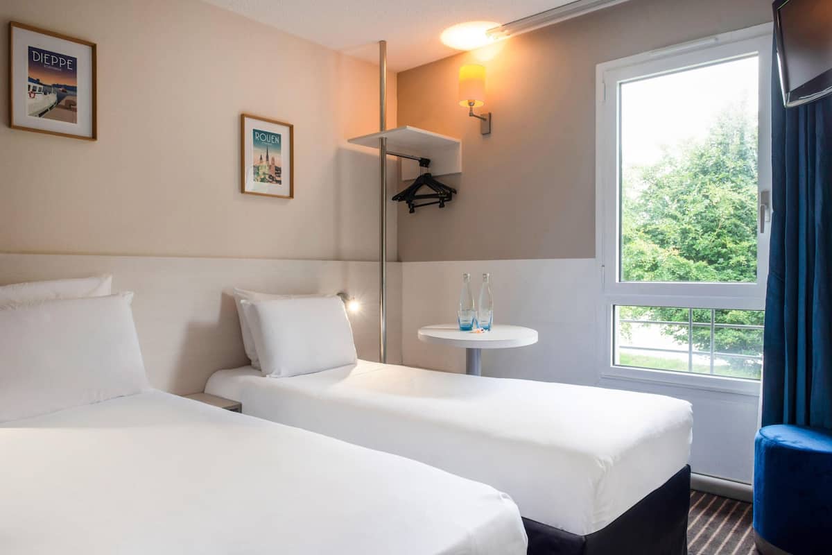 A hotel room with two beds, a small table with bottles, a wall-mounted lamp, and framed pictures on the wall.
