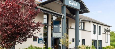 Un edificio llamado 'Kyriad' con un estilo arquitectónico moderno, que presenta una entrada destacada, un área de estar al aire libre y jardines bien cuidados.
