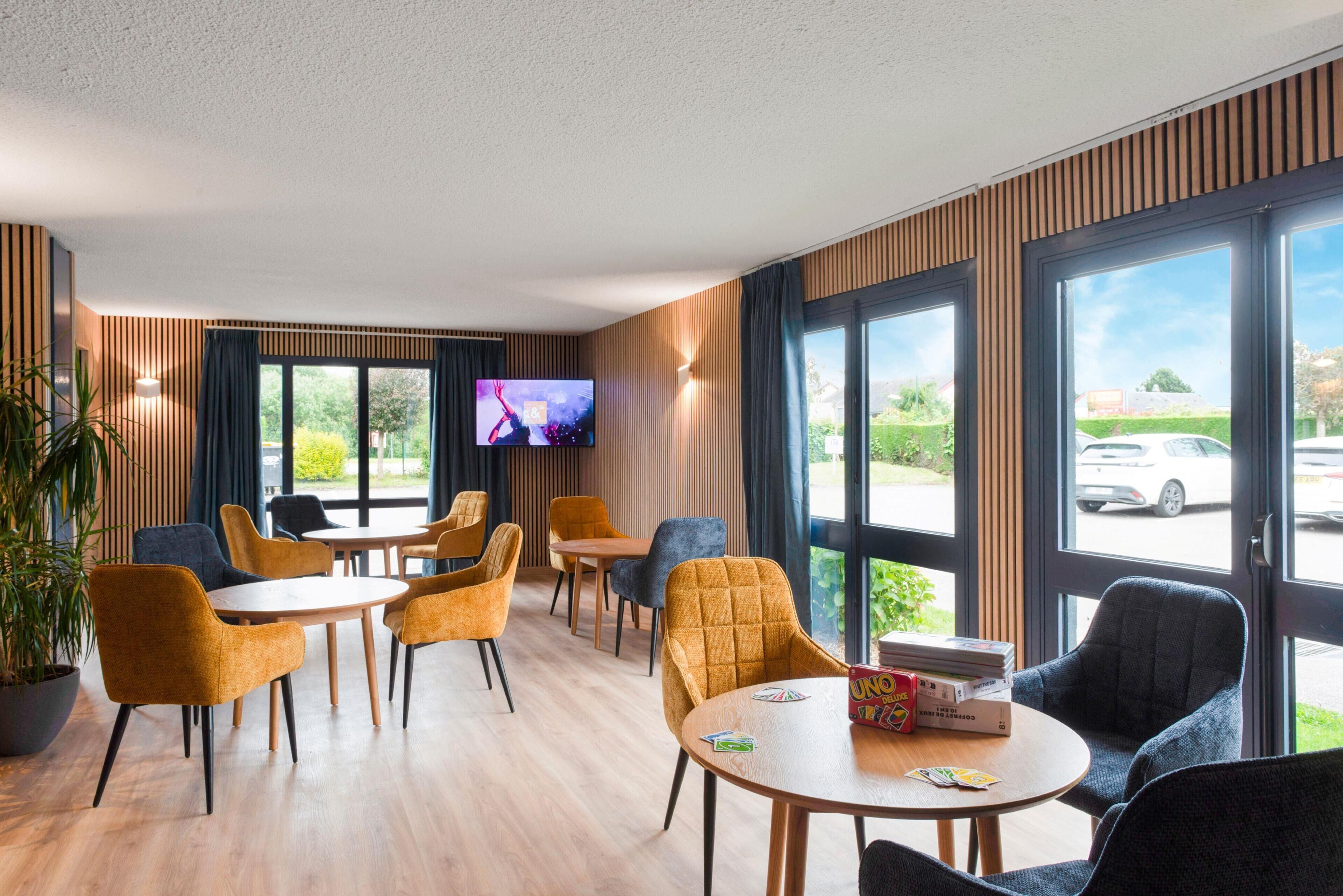 Un hall d’hôtel moderne avec un revêtement de sol en bois, divers types de sièges, notamment des tables et des chaises rondes, un téléviseur à écran plat fixé au mur et de grandes fenêtres offrant une vue sur l’extérieur.