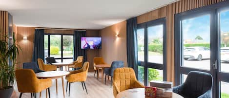Un hall d’hôtel moderne avec un revêtement de sol en bois, divers types de sièges, notamment des tables et des chaises rondes, un téléviseur à écran plat fixé au mur et de grandes fenêtres offrant une vue sur l’extérieur.