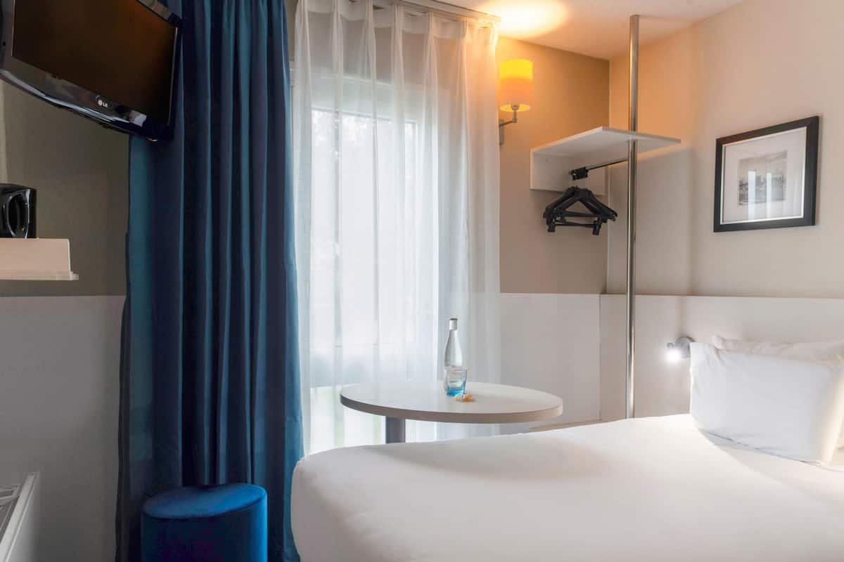 A hotel room with a bed, a small round table, a TV mounted on the wall, a coat rack, and a framed picture on the wall.
