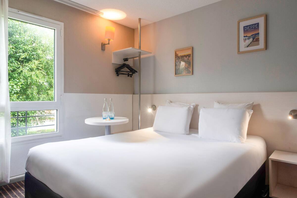 A hotel room with a large bed, a small round table with a bottle, a wall-mounted lamp, and a framed picture on the wall.