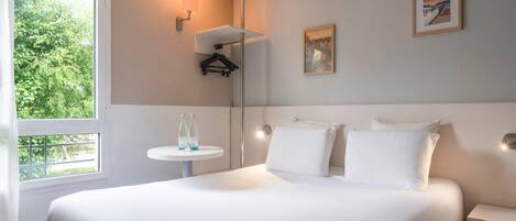 A hotel room with a large bed, a small round table with a bottle, a wall-mounted lamp, and a framed picture on the wall.