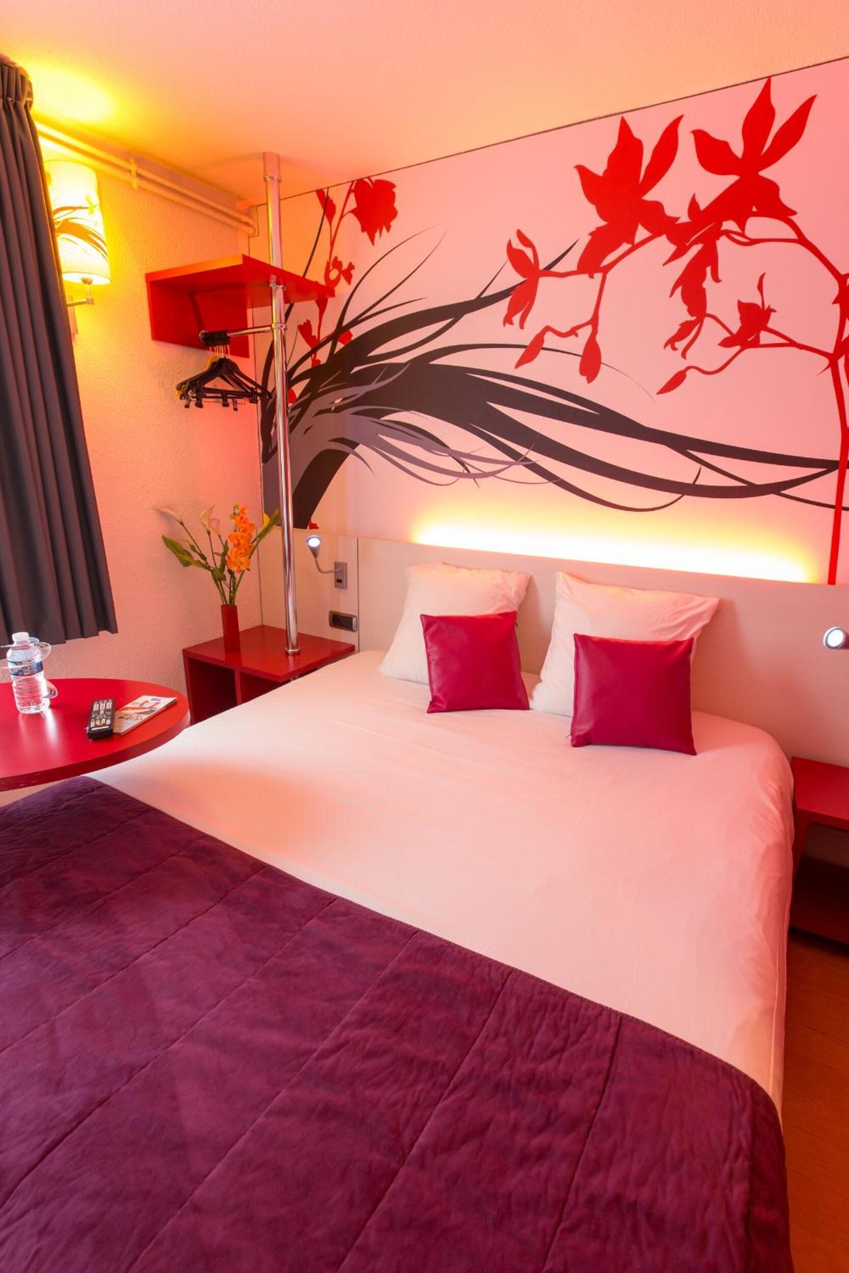 Hotelkamer met een groot bed, rode kussens, een rode nachtkastje, een rode tafel met een waterfles en een afstandsbediening, en een wand met een rood bloemenmotief.