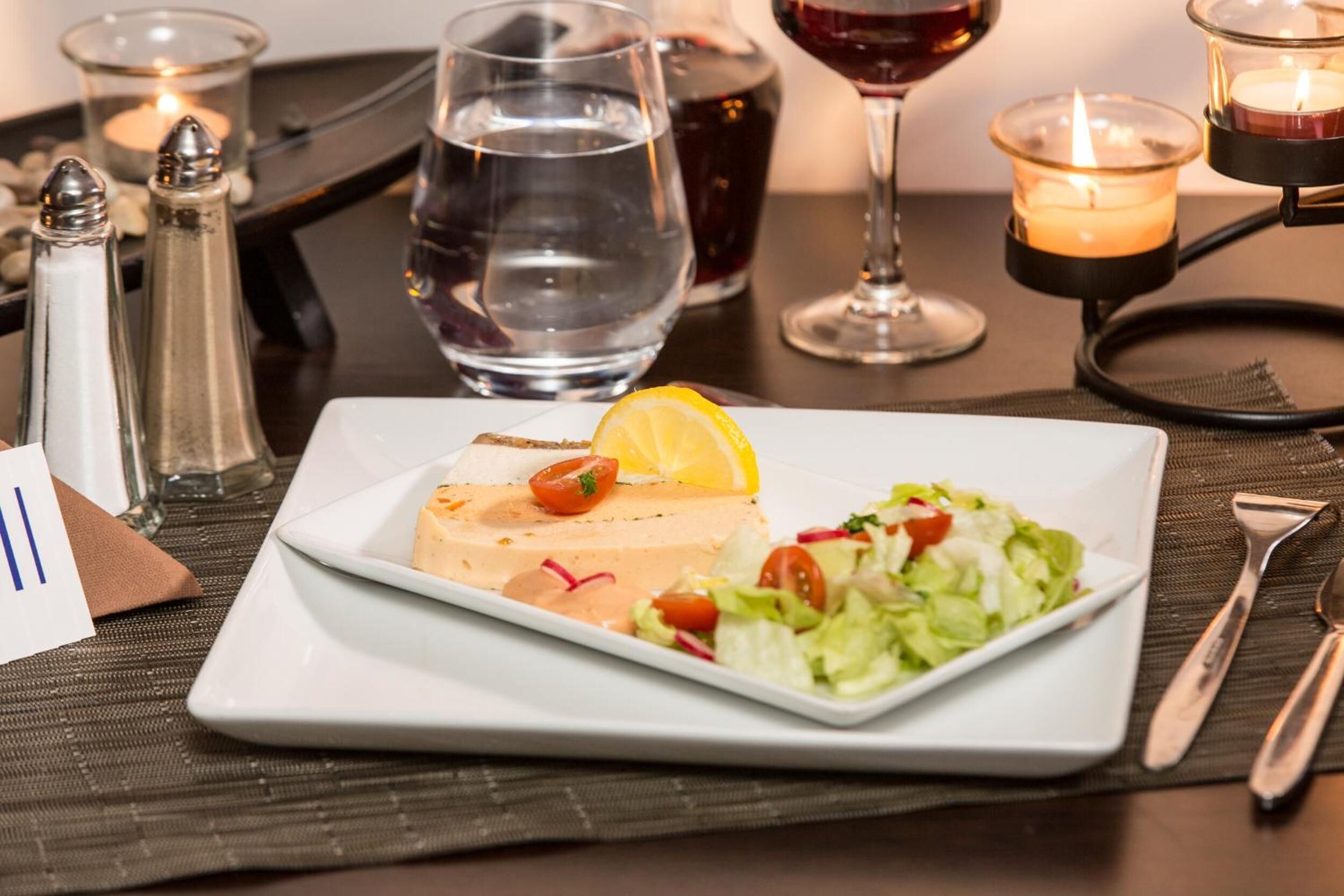 A white plate with a slice of salmon, lemon, and salad on a table with candles, salt and pepper shakers, and a glass of water.