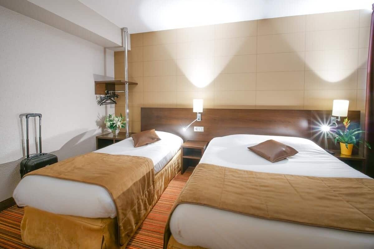 A hotel room with two single beds, a small table, a chair, a suitcase, and a potted plant.