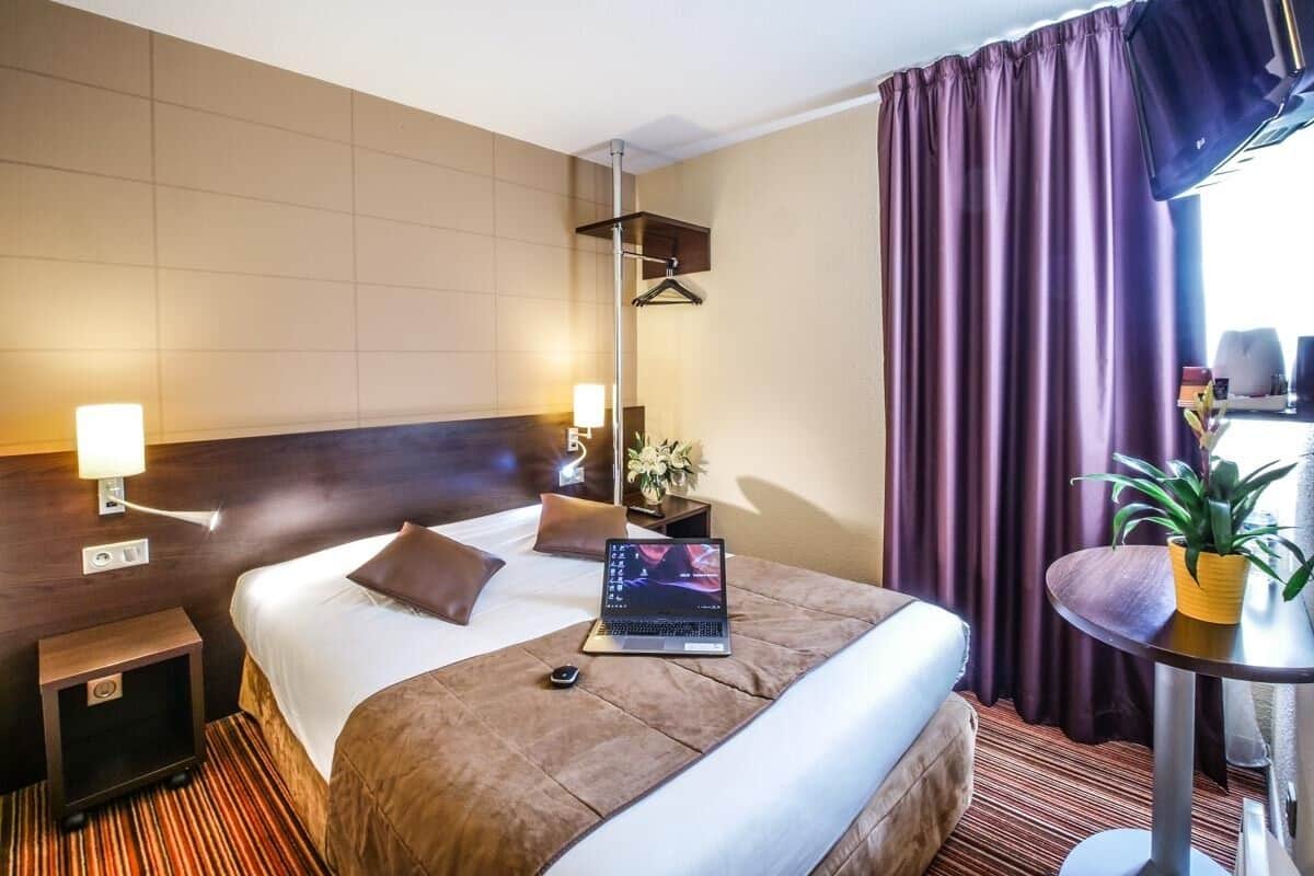 A hotel room with a bed, bedside table, lamp, laptop, and a potted plant.