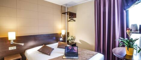 Una habitación de hotel con una cama, mesita de noche, lámpara, laptop y una planta en maceta.