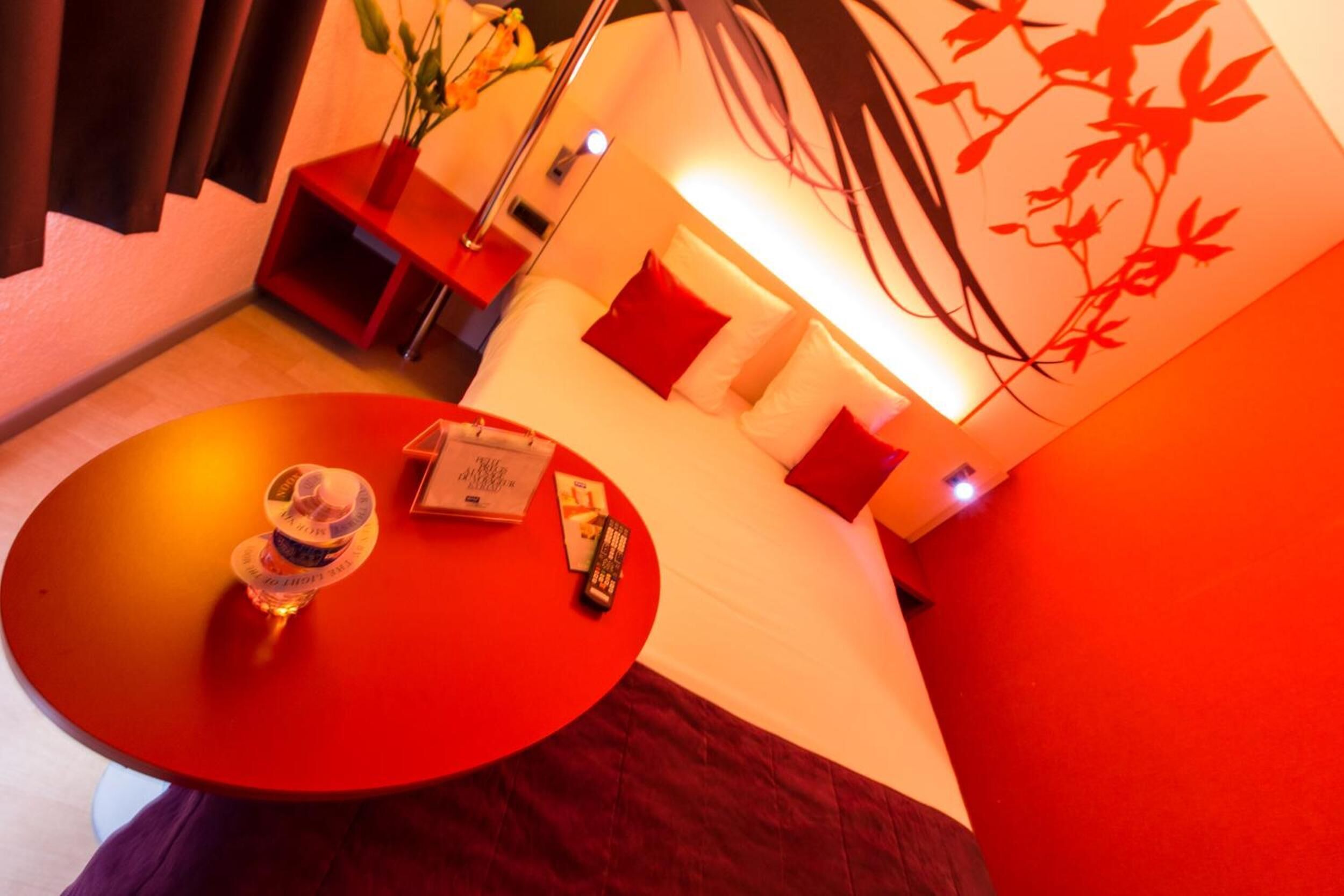A hotel room with a red table, a bed with red pillows, a bedside table with a lamp, and a wall with a red floral design.