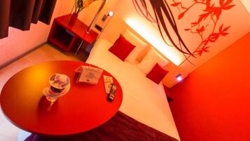 Habitación de hotel con una mesa roja, una cama con almohadillas rojas, una mesita de noche con una lámpara y una pared con diseño floral rojo.