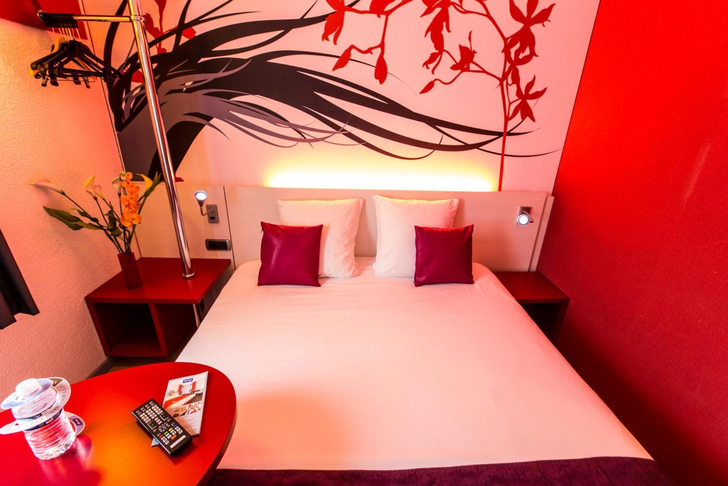 A hotel room with a large bed, red and white pillows, a nightstand with a lamp, a small table with a remote control, and a vase with flowers.