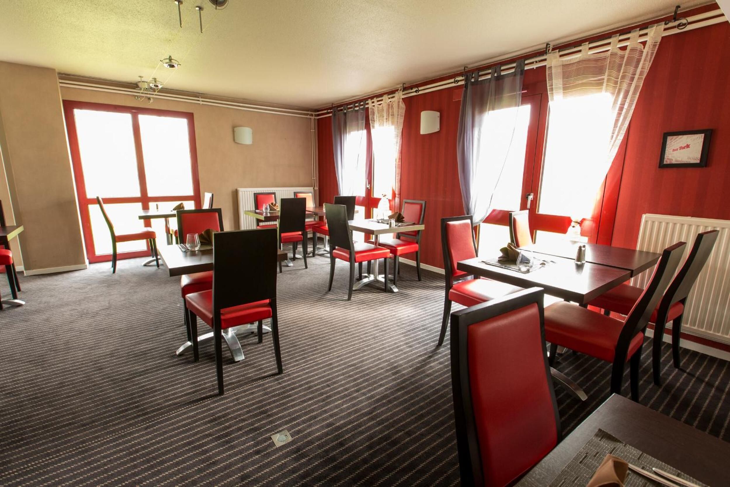 A hotel room with a dining area featuring red chairs and tables, large windows with curtains, and a carpeted floor.