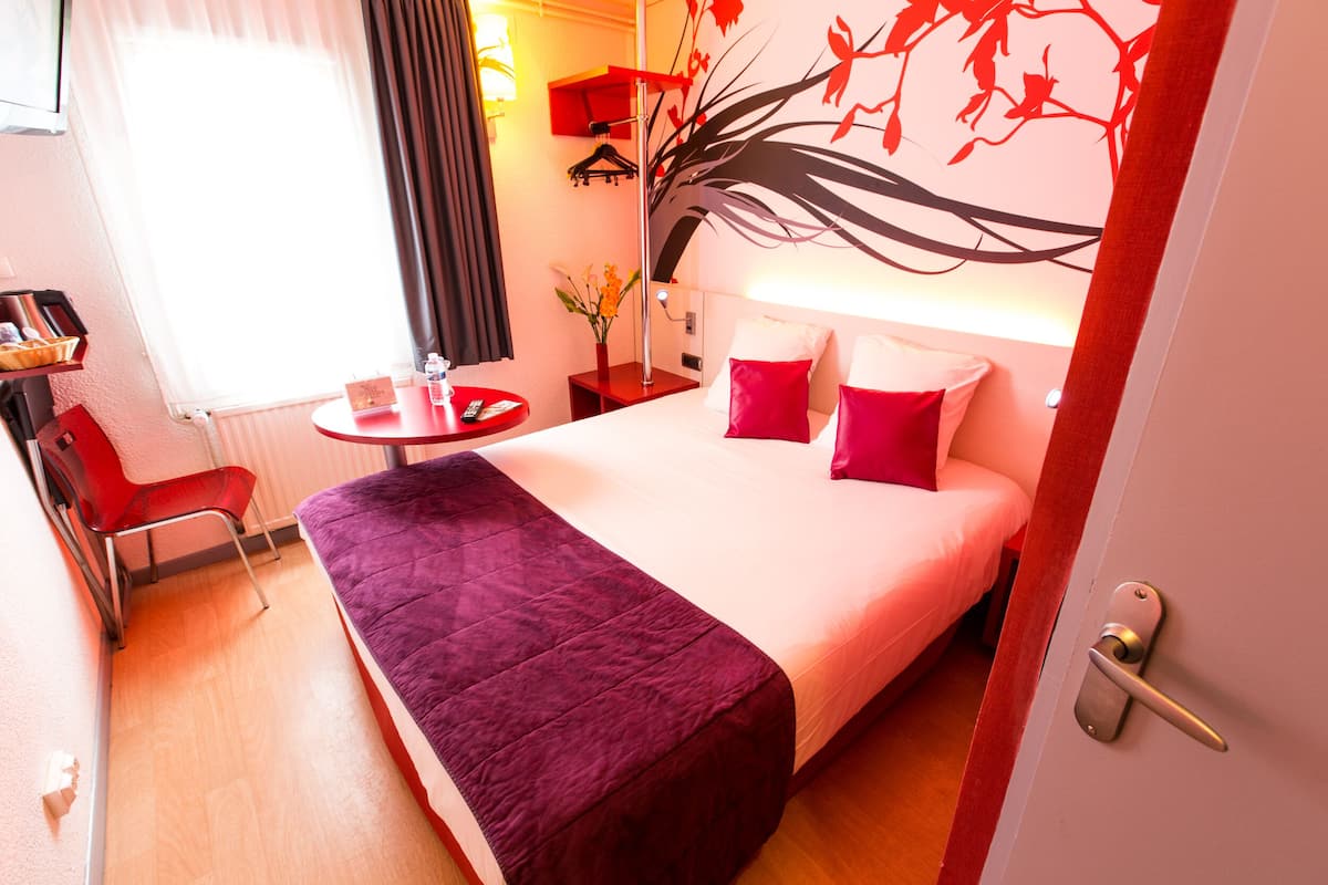 A hotel room with a bed, a chair, a small table, and a wall with a tree mural.