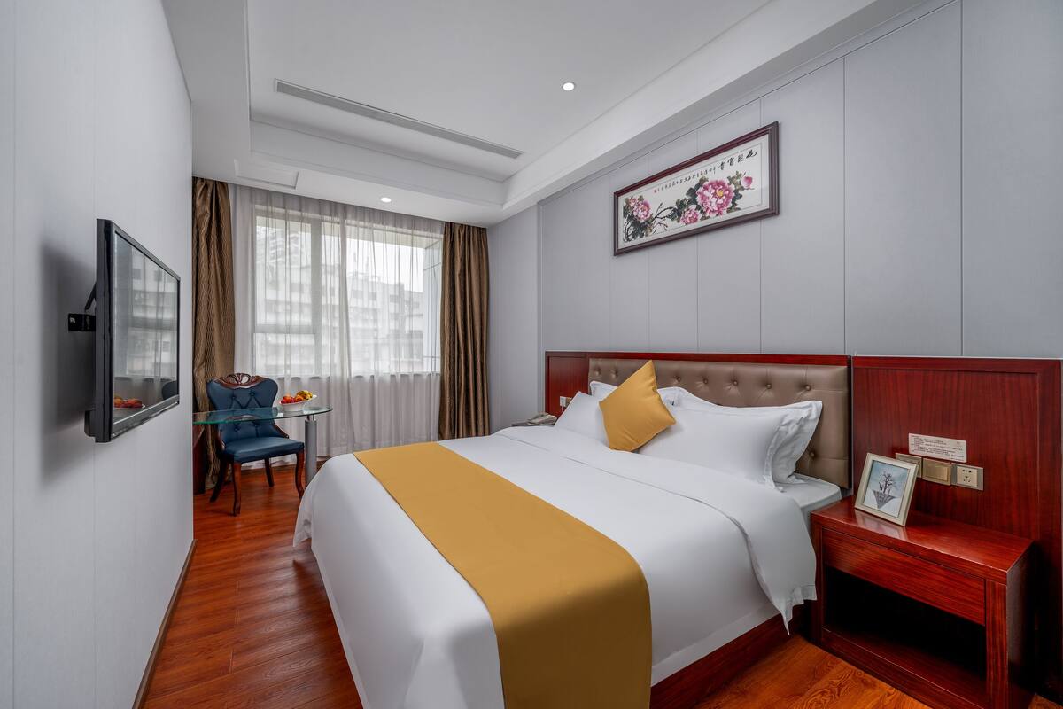 Een hotelkamer met een groot bed, een nachtkastje met een fotolijstje, een aan de muur bevestigde televisie en een raam met gordijnen.