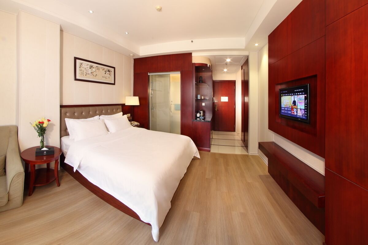 A hotel room with a large bed, a TV mounted on the wall, a small table with a vase of flowers, and a wooden wardrobe.