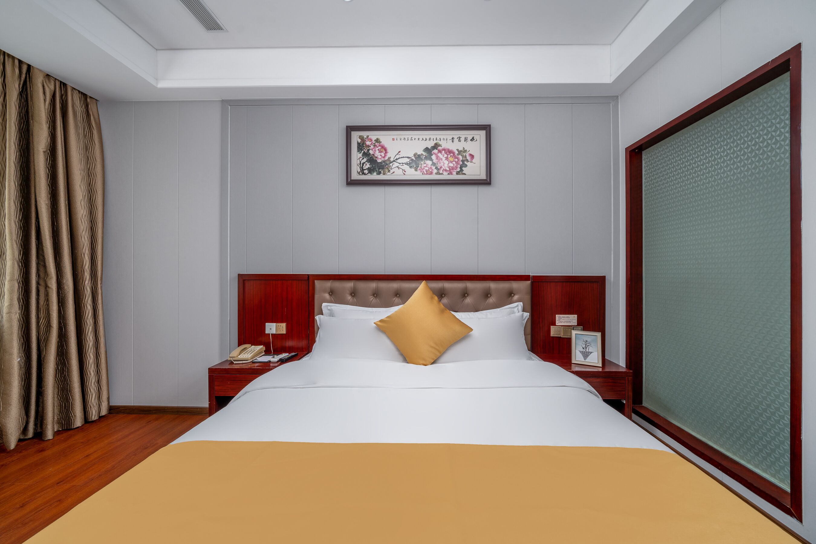 A hotel room with a large bed, a wooden headboard, a nightstand with a telephone, and a framed artwork on the wall.
