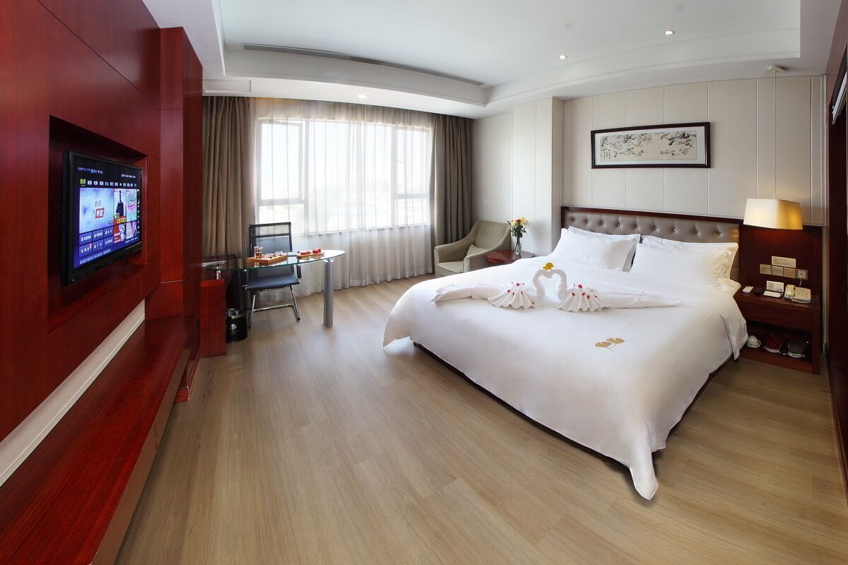 A hotel room with a large bed, a TV mounted on the wall, a desk with a chair, and a window with curtains.