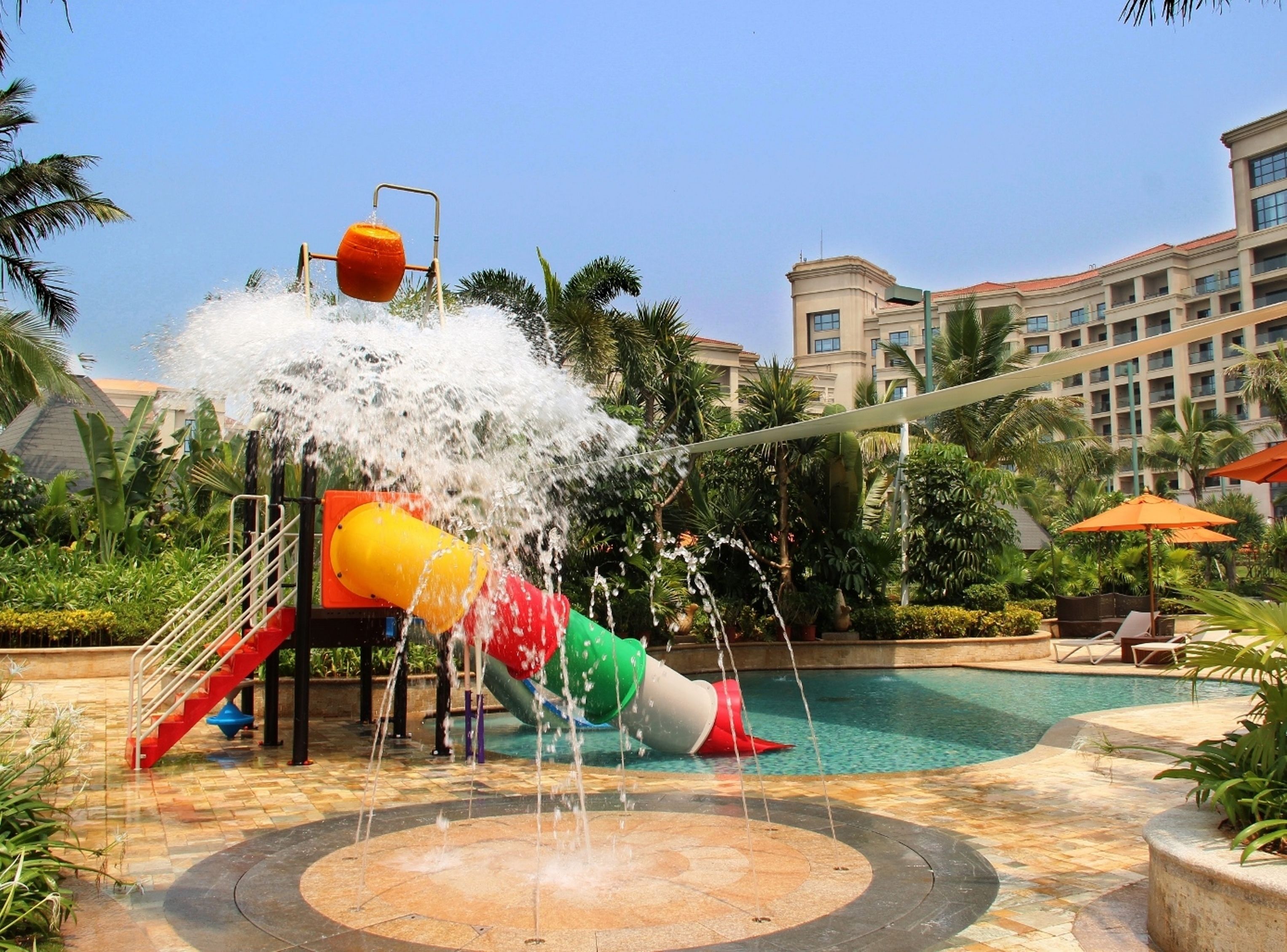 A water play area with a slide and water jets, surrounded by a pool and palm trees, with a building in the background.
