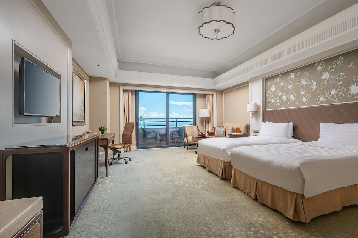 A hotel room with a large bed, a desk with a chair, a television, and a view of the ocean through a sliding door.