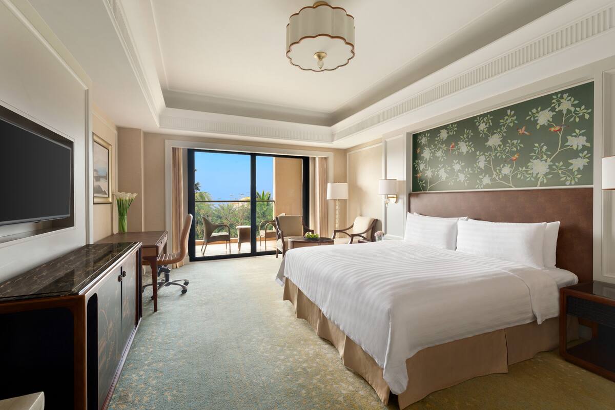 A hotel room with a large bed, a desk, a TV, a balcony with a view, and a floral wall decoration.