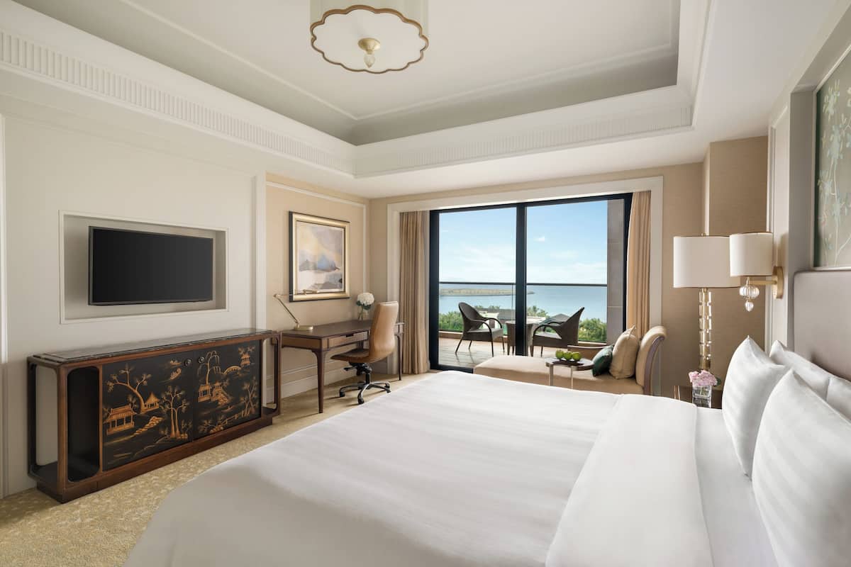 A hotel room with a large bed, a desk with a chair, a television, and a balcony with a view.