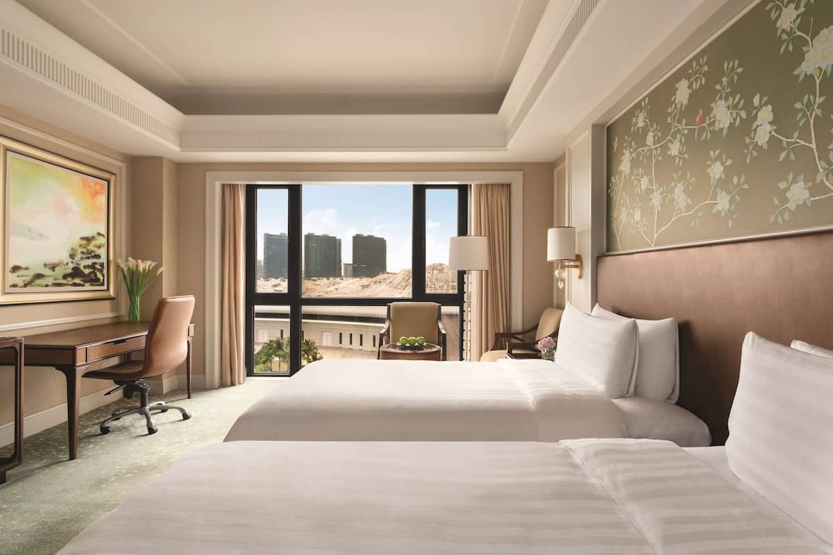 A hotel room with a large bed, a desk, and a chair. There is a view of buildings outside the window.