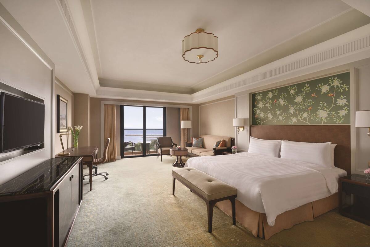 A hotel room with a large bed, a desk with a TV, a chair, a bench, and a view of the ocean.
