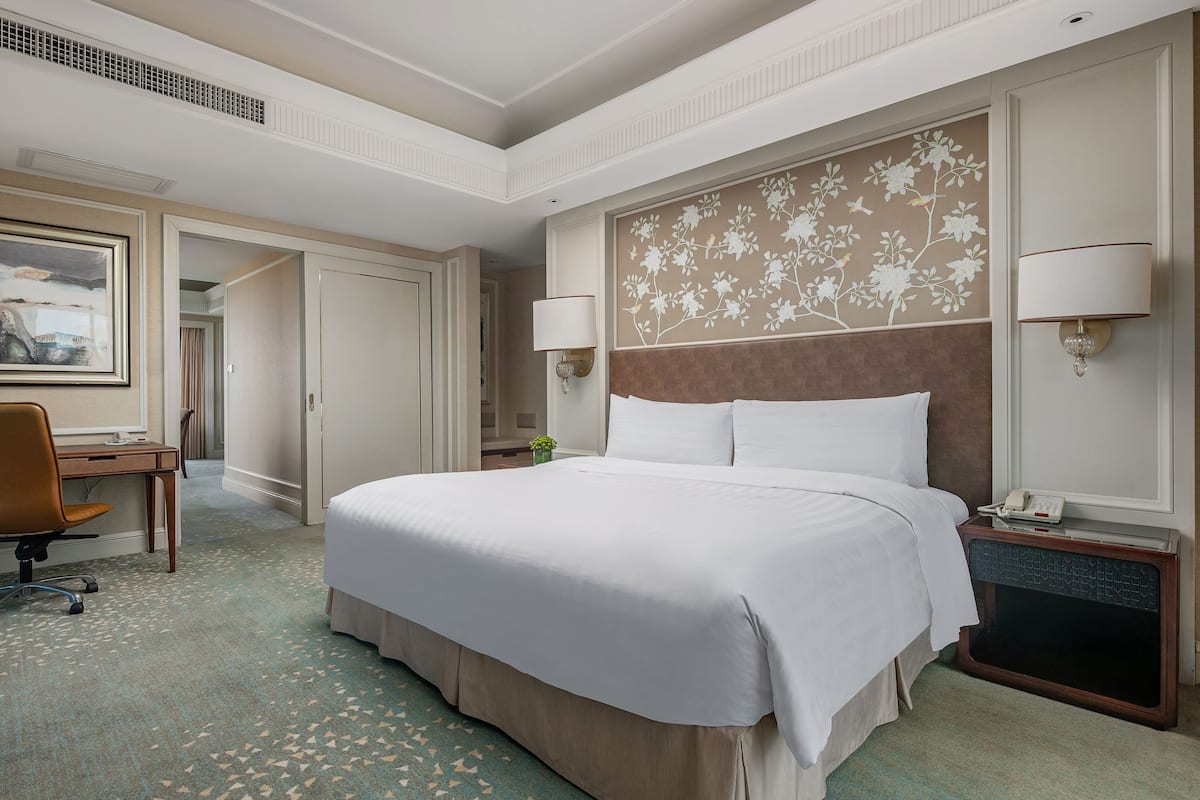 A hotel room with a large bed, a desk, a chair, and a nightstand with a lamp.