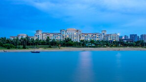Un gran hotel con varios pisos, una playa en primer plano y palmeras.