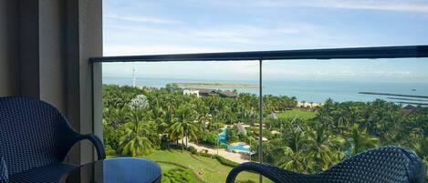 A balcony with wicker chairs and a glass table overlooking a resort with a pool, palm trees, and a view of the sea.