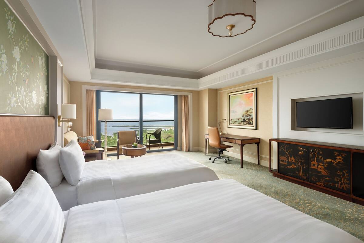 A hotel room with two beds, a desk, a chair, a TV, and a large window offering a view.