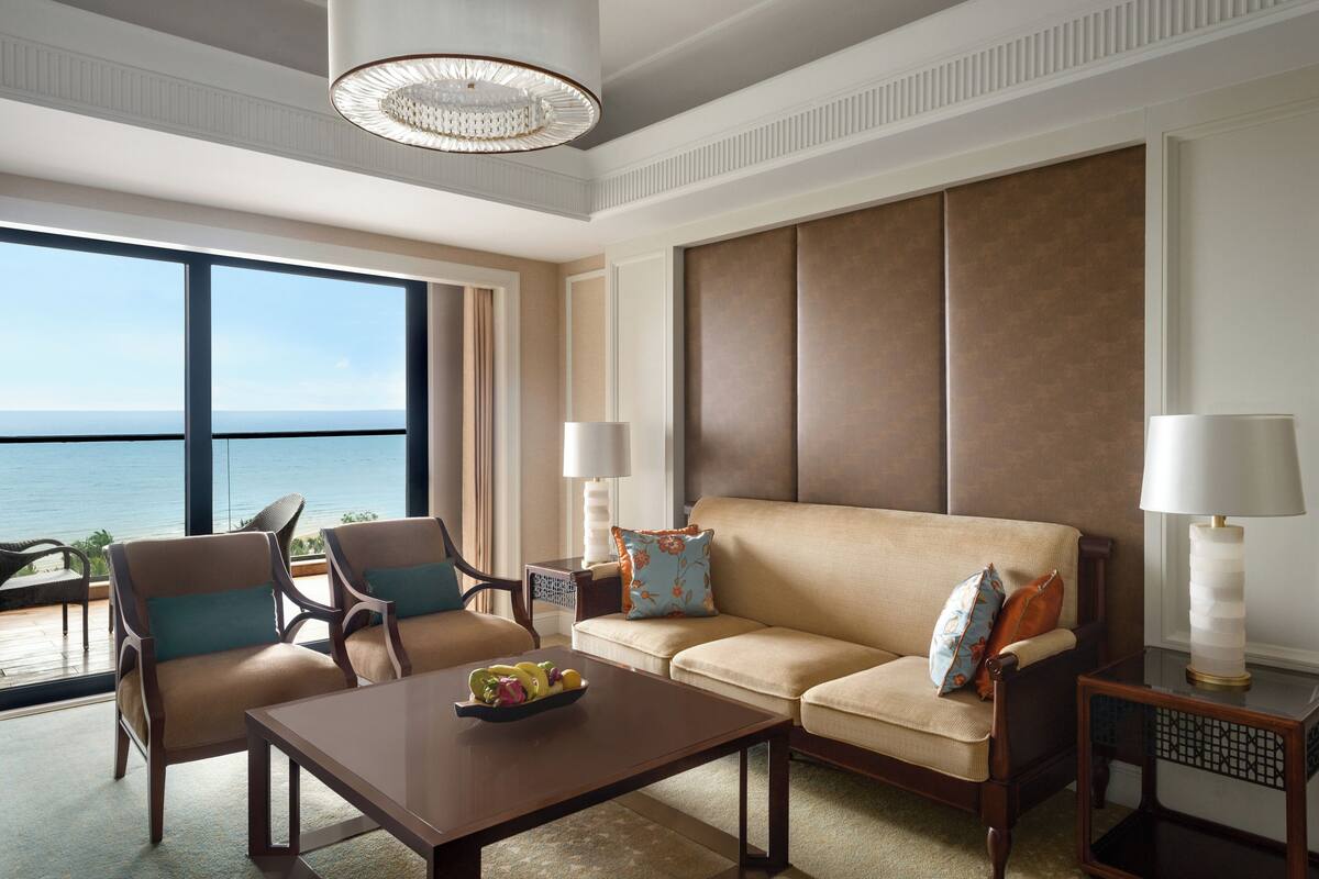 A spacious living room with a large window offering a view of the sea, a beige sofa, two armchairs, a coffee table with fruit, and two side tables with lamps.