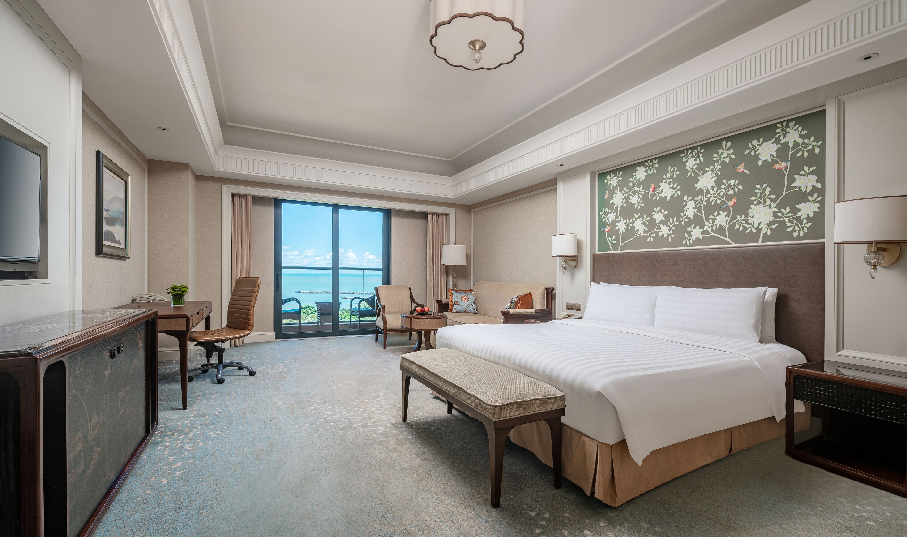 A hotel room with a large bed, a desk, a chair, a TV, and a view of the ocean.