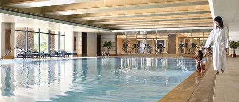 An indoor swimming pool with clear water, a wooden ceiling, and a person and child near the edge.